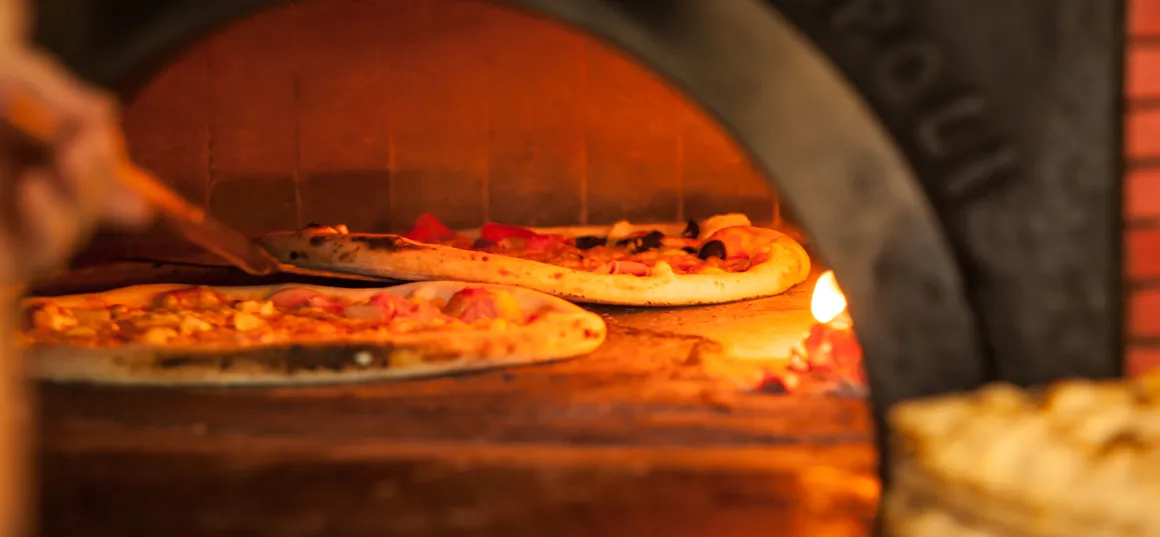Deux pizzas sur une pelle devant la bouche d’un four à briques allumé